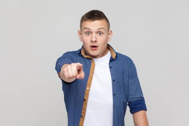 Portrait of astonished amazed handsome teenager boy wearing blue shirt pointing to camera with surprised shocked expression, making choice. Indoor studio shot isolated on gray background.