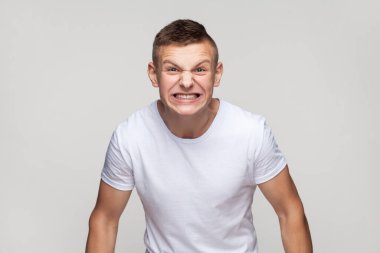 Portrait of handsome young teenager boy wearing T-shirt expressing aggressive emotions, screaming with hate and anger, frowning face. Indoor studio shot isolated on gray background.