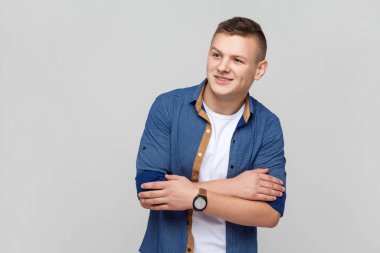 Portrait of handsome attractive young smiling teenager boy wearing blue shirt keeps hands folded, looking away with dreaming expression. Indoor studio shot isolated on gray background.