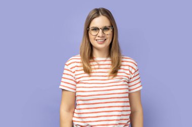 Portrait of smiling optimistic blond woman wearing striped T-shirt standing in optical glasses and looking at camera with satisfied look. Indoor studio shot isolated on purple background.