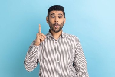 Portrait of inspired handsome bearded businessman pointing finger up and looking amazed about sudden genius idea, got solution, wearing striped shirt. Indoor studio shot isolated on blue background.