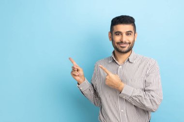 Satisfied bearded businessman pointing aside, showing blank copy space for idea presentation, commercial text, smiling happily, wearing striped shirt. Indoor studio shot isolated on blue background.