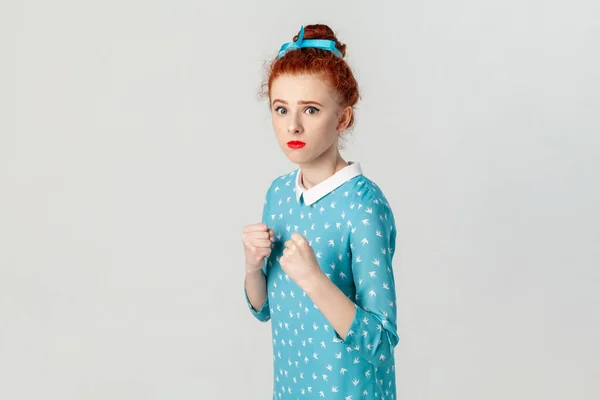 Portrait of confident serious red haired woman with hair bun, standing with clenched fists, being ready to attack, boxing, wearing blue dress. Indoor studio shot isolated on gray background.
