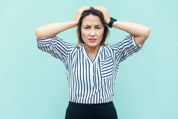 Portrait of unhealthy sick middle aged woman wearing striped shirt standing with raised hands, suffering terrible headache, feels unwell. Indoor studio shot isolated on light blue background.