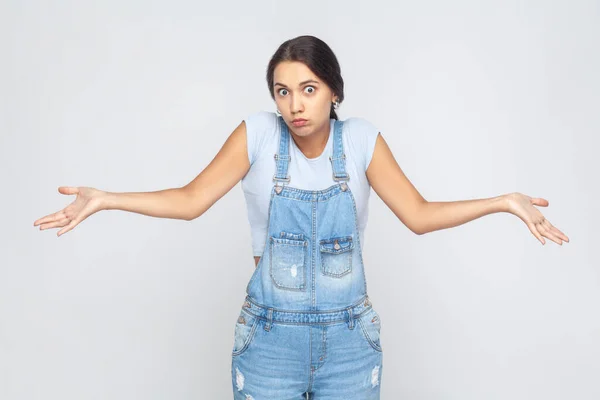 Maybe, don\'t know. Portrait of unsure woman wearing denim overalls raising hands with confused perplexed puzzled expression, not sure about decision. Indoor studio shot isolated on gray background.