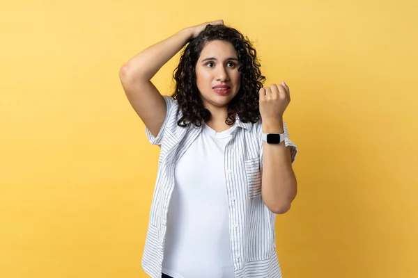 Portrait of beautiful adorable confused woman with dark wavy hair standing with puzzled facial expression, looks pensive, showing smart watch. Indoor studio shot isolated on yellow background.