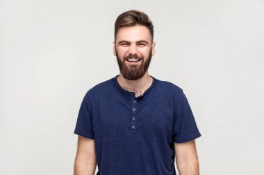 Portrait of overjoyed extremely happy handsome man with beard wearing dark blue T-shirt hunching from laughter, laughing out loud, crazy face. Indoor shot isolated on gray background.
