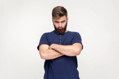Portrait of man with beard wearing dark blue T-shirt being upset of bad news, looking at camera with frowning face, expressing sadness, keeps arms crossed. Indoor shot isolated on gray background.