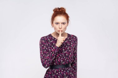 Please, be quiet. Portrait of redhead woman wearing dress holding finger on lips making hush silence gesture, asking to keep secret, don't speak. Indoor studio shot isolated on gray background.
