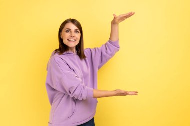 Portrait of happy charming woman presenting copy space between hands, holding empty place for commercial text, wearing purple hoodie. Indoor studio shot isolated on yellow background.