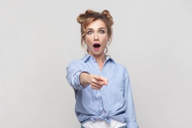 Startled emotional blonde woman points directly at camera with index finger, opens mouth from amazement, has eyes popped out, wearing blue shirt. Indoor studio shot isolated on gray background.