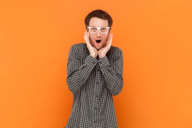 Amazed man nerd doesn`t believe his success, keeps hands on head, stares at camera, says omg or wow, wearing shirt with blue bow tie and white glasses. Indoor studio shot isolated on orange background