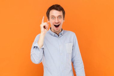 Portrait of inspired surprised man standing with raised finger, having sudden smart idea, looking at camera with open mouth, wearing light blue shirt. Indoor studio shot isolated on orange background.