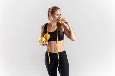 Portrait of attractive blonde woman standing with tape measure, holding fruit in hands, drinking fresh orange juice, wearing black fitness clothing. Indoor studio shot isolated on gray background.