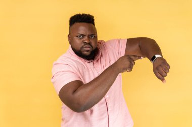 Time is out. Portrait of serious handsome bearded man wearing pink shirt standing and looking at camera, pointing on his smart watch. Indoor studio shot isolated on yellow background.