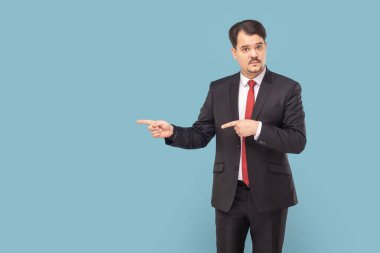 Serious strict man with mustache standing pointing aside at advertisement area, copy space for promotion, wearing black suit with red tie. Indoor studio shot isolated on light blue background.