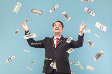 Oh my god, wow. Portrait of man with mustache absolutely shocked of money rain falling from up, rejoicing, wearing black suit with red tie. Indoor studio shot isolated on light blue background.