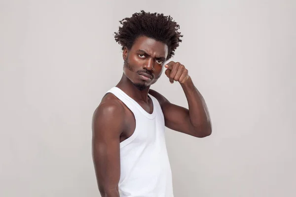 Portrait of serious strict angry man with Afro hairstyle points index finger at camera warns you about something, choosing you, wearing white T-shirt. Indoor studio shot isolated on gray background.