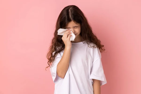 Portrait of sad upset unhappy little girl wearing white T-shirt crying, hard going through loss or defeat, feeling sorrow, holding handkerchief. Indoor studio shot isolated on pink background.