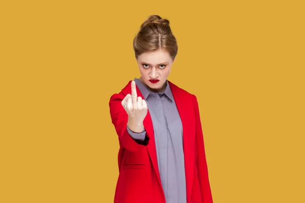 Portrait of aggressive rude woman with red lips standing showing middle finger to camera, arguing with somebody, wearing red jacket. Indoor studio shot isolated on yellow background.