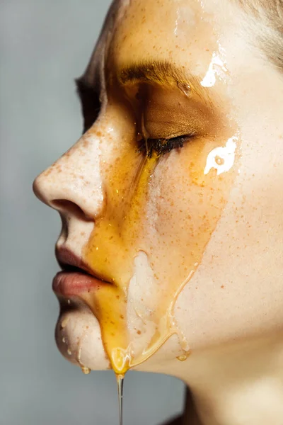 Closeup side view portrait of beautiful serious young brunette woman with freckles, applying honey on face for beauty treatment. Indoor studio shot isolated on gray background.