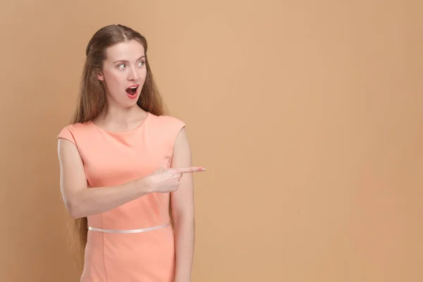 Portrait of shocked amazed surprised woman with long hair pointing aside at advertisement area, copy space for promotion, wearing elegant dress. Indoor studio shot isolated on brown background.