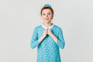 Wow. Portrait of amazed excited beautiful ginger woman being surprised, sees something amazing, keeps hands together, wearing blue dress. Indoor studio shot isolated on gray background.