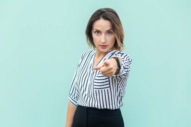 Portrait of angry serious middle aged woman wearing striped shirt standing pointing to camera, choosing you, looking at bossy expression. Indoor studio shot isolated on light blue background.