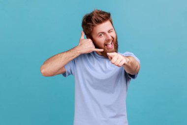 You call me. Portrait of playful bearded man flirting and pointing at camera with attractive smile, making call me gesture with fingers. Indoor studio shot isolated on blue background.