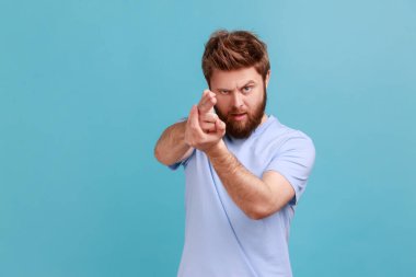 Portrait of focused bearded man pointing finger gun gesture to target, threatening to kill, shooting with hand pistol and looking menace. Indoor studio shot isolated on blue background.