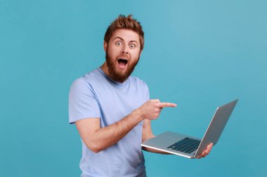 Portrait of shocked bearded man pointing finger at laptop and looking at camera with opened mouth and big eyes, expressing astonishment. Indoor studio shot isolated on blue background.