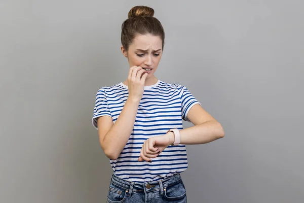 I need more time. Portrait of nervous woman wearing striped T-shirt standing and looking at her smart watch and want more time, biting fingernails. Indoor studio shot isolated on gray background.