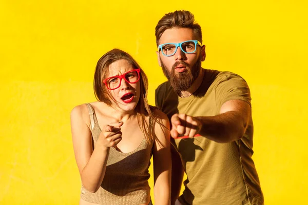 Portrait of astonished amazed man and woman in glasses standing with shocked face and pointing to camera, having surprised expression. Indoor studio shot isolated on yellow background.