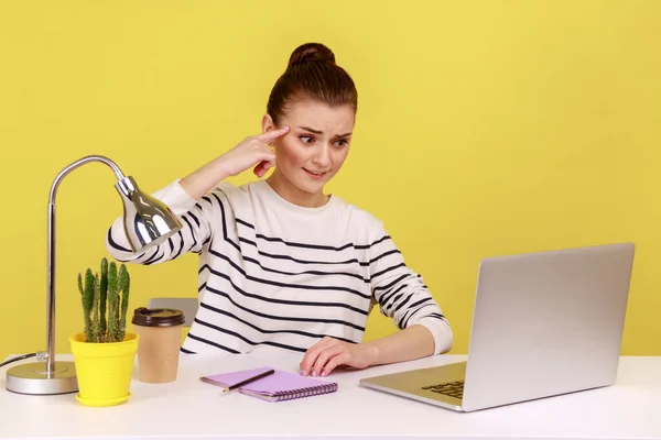 办公室女职员坐在工作场所 用手指对着笔记本电脑屏幕 表现出愚蠢的姿态 脸上带着不愉快的疲倦表情 在黄色背景下隔离的室内工作室拍摄 — 图库照片