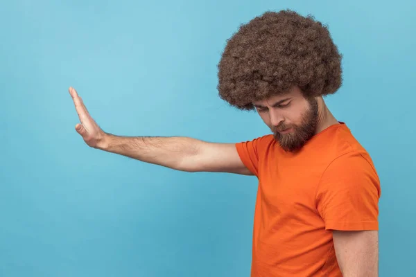 Side view of man with Afro hairstyle in T-shirt making stop gesture showing palm of hand, conflict prohibition warning about danger, stop bullying. Indoor studio shot isolated on blue background.