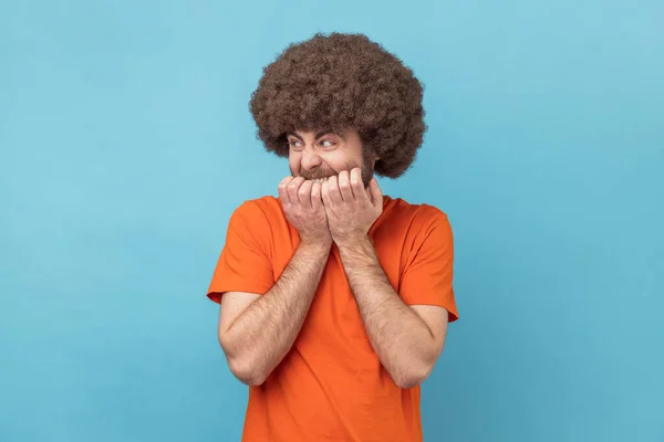 Anxiety disorder. Portrait of man with Afro hairstyle wearing orange T-shirt biting nails, nervous about troubles, panicking and looking scared. Indoor studio shot isolated on blue background.