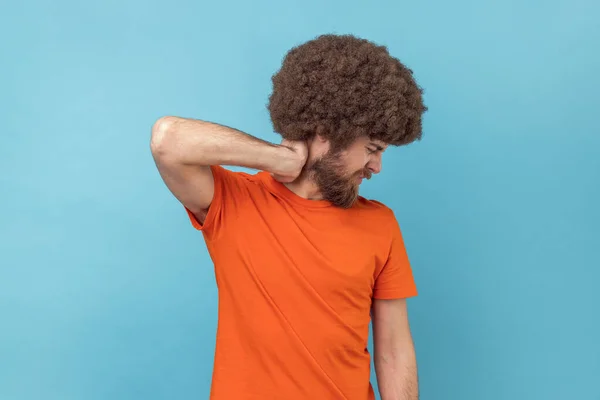 Portrait of man with Afro hairstyle in T-shirt touching neck, feeling acute pain moving and turning head, suffering spine problems, osteochondrosis. Indoor studio shot isolated on blue background.