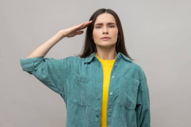 Yes sir. Serious smart young woman giving salute, listening carefully to order, looking with responsible attentive expression, wearing casual jacket. Indoor studio shot isolated on gray background.