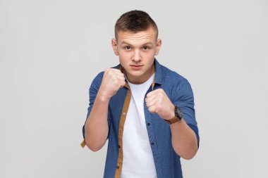 Portrait of teenager boy in blue shirt standing with boxing fists and ready to attack or defence, looking with angry face, looking at camera with aggression. Indoor studio shot isolated on gray background
