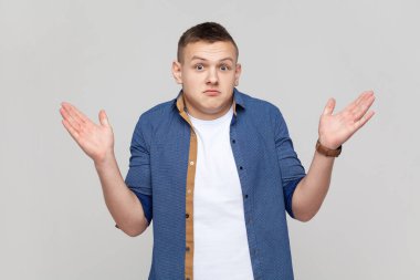 I don't know, who cares. Portrait of confused clueless teenager boy wearing blue shirt shrugging shoulders, making no idea gesture, whatever. Indoor studio shot isolated on gray background.