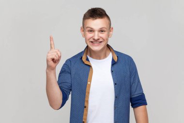 Portrait of excited amazed teenager boy wearing blue shirt pointing finger up with genius idea, surprised by suddenly invented smart solution. Indoor studio shot isolated on gray background.