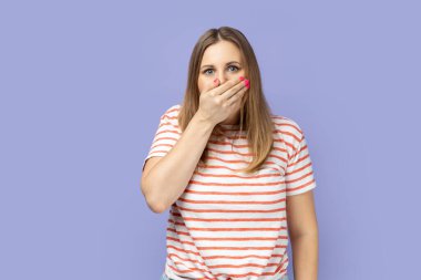 Şaşkın sarışın kadın portresi çizgili tişört giyiyor ağzını avuç içiyle kapatıyor, kimseye bir şey söyleme. Mor arka planda izole edilmiş kapalı stüdyo çekimi.
