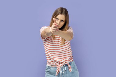 Portrait of serious brave blond woman wearing striped T-shirt holding fingers pretending holding gun, aiming enemy, self-defense, looking at camera. Indoor studio shot isolated on purple background.