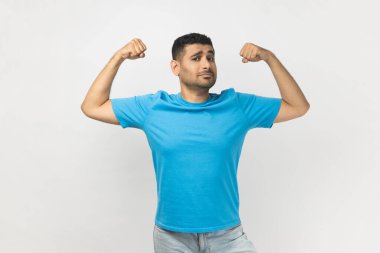 Portrait of confident strong powerful unshaven man wearing blue T- shirt standing raised his arms, showing his biceps and power. Indoor studio shot isolated on gray background.