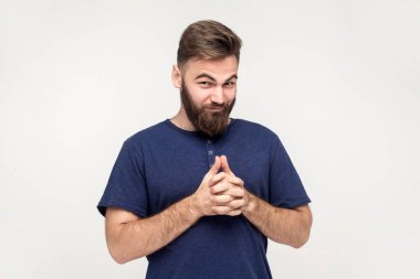 Portrait of devious cunning man with beard wearing dark blue T-shirt clasping hands and smirking mysteriously, scheming cheats, evil prank. Indoor shot isolated on gray background.