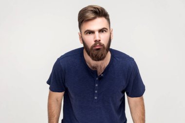 Portrait of disgruntled man with beard wearing dark blue T-shirt looking with annoyed indignant expression at camera, being angry and aggressive. Indoor shot isolated on gray background.