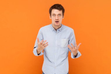 Portrait of shocked unhappy young adult man standing with raised hands, asking what, misunderstanding, wearing light blue shirt. Indoor studio shot isolated on orange background.