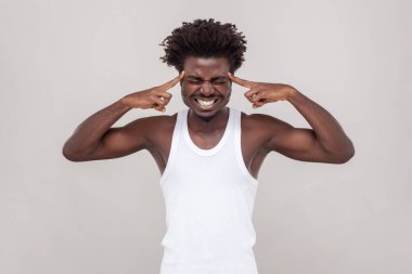 Portrait of unhealthy sick man with Afro hairstyle touches temples has unbearable headache suffers from migraine, wearing white T-shirt. Indoor studio shot isolated on gray background.