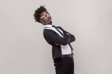 Side view portrait of funny man in glasses standing with crossed arms, looking away with confused puzzled expression, wearing white shirt and tuxedo. Indoor studio shot isolated on gray background.