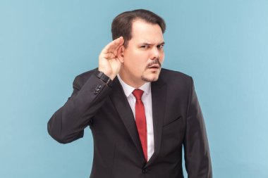 Portrait of concentrated curious man with mustache standing with hand near ear, listening private information, wearing black suit with red tie. Indoor studio shot isolated on light blue background.